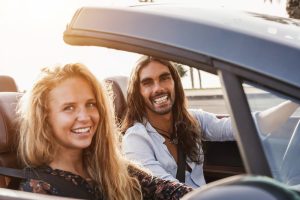 A man and a woman driving on a California highway in a convertible. The sun is setting in the foreground and a palm tree can be spotted nearby.